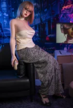 Elena, a 165cm gamer girl silicone doll, sits beside a monitor in rectangular glasses, a cream halter top, and gray patterned pants while holding a game controller.