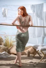 Facing the sea at the bamboo rail, Faye shows the lace-tied open back of her sage-green dress and turns her head to the side.