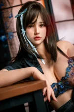 Mika, a 161cm realistic companion doll, seated beside the wooden table with one arm on the edge, wearing a blue floral lace bra.