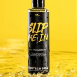 An upright bottle of Slip Me In lubricant stands in shallow splashing water against a yellow background.