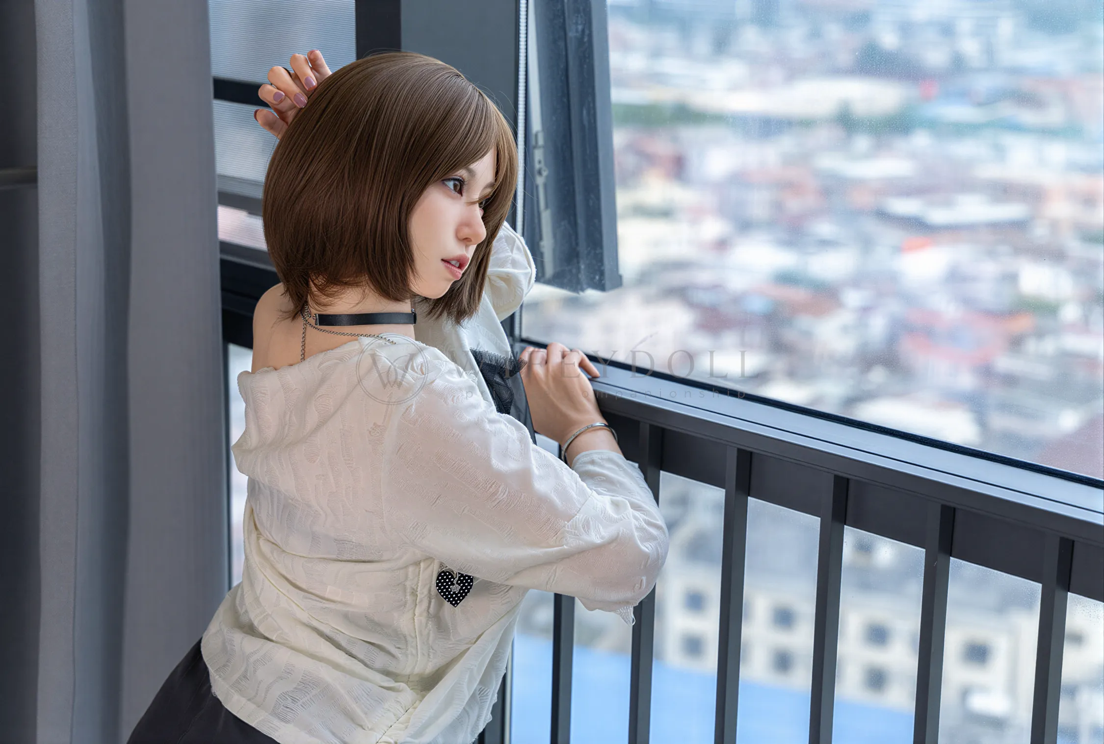 Suki stands by the balcony railing in her loose white sweater, turning her short brown bob toward the city view.