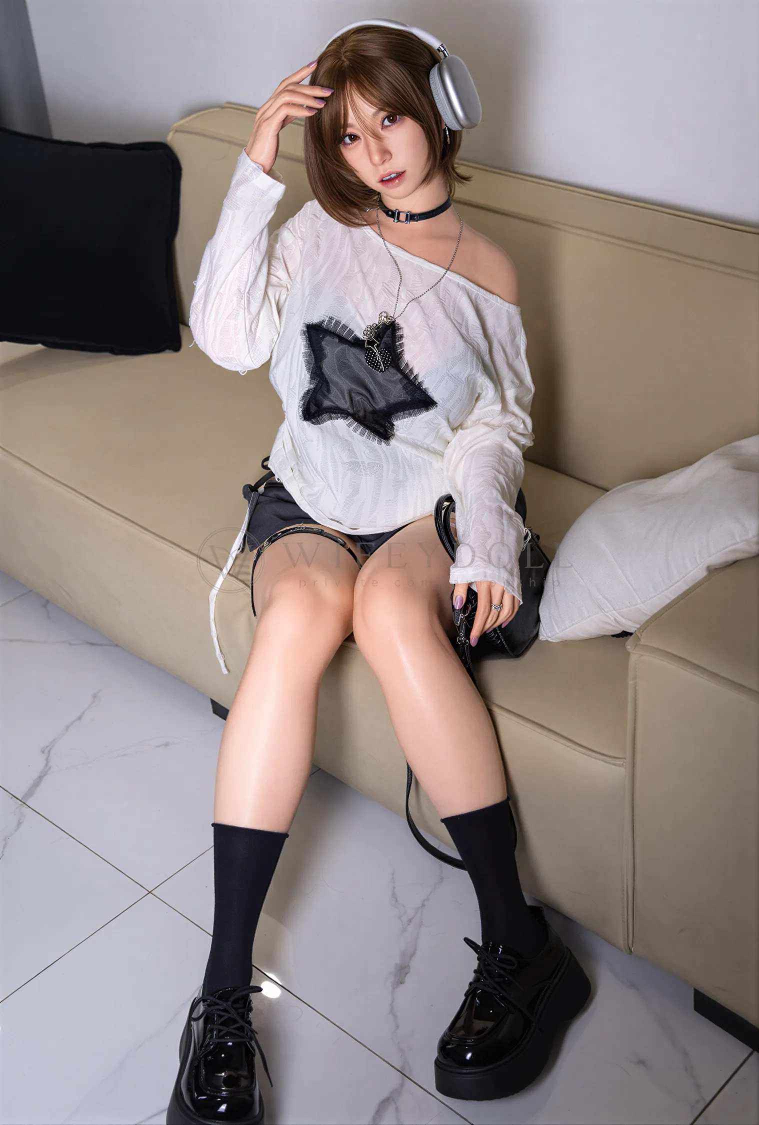 Suki, a 165cm silicone sex doll, sits at the edge of a beige sofa with silver headphones on, her loose white star sweater slipping off one shoulder.