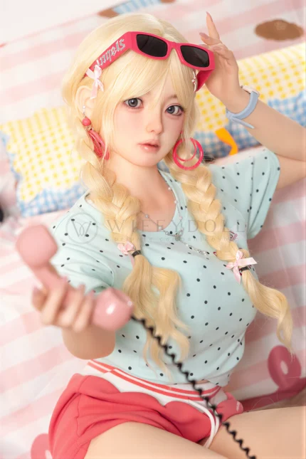 Xia Ying silicone companion doll seated on the bed, holding a pink phone handset and reaching one hand forward.