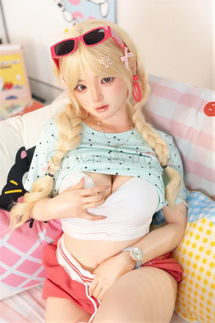 Xia Ying silicone doll seated on the bed in a white top, one hand resting over her chest.