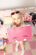 Xia Ying silicone doll seated on the bed holding a large pink card in front of her legs.