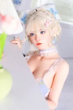 Zia, a 154cm realistic sex doll, stands beside the ribbon-trimmed shelf in a pale blue gingham outfit with blonde braids.