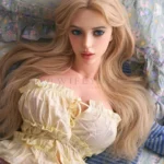 Closer portrait of Bailey in a pale yellow ruffled top with long blonde hair spread across the bedding, presenting Bailey as a Bailey silicone doll.