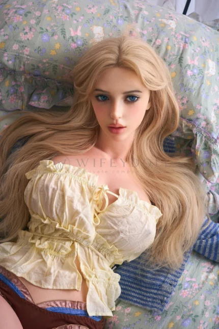 Closer portrait of Bailey in a pale yellow ruffled top with long blonde hair spread across the bedding, presenting Bailey as a Bailey silicone doll.