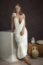Nina silicone sex doll standing in a Greek-style white dress and tan lace-up sandals.