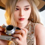 Vivienne realistic silicone sex doll facing forward in a straw hat, holding a camera over her floral-trimmed top.